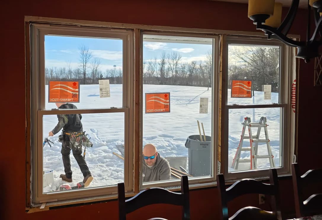Two workers are seen outside in the snow installing windows on a house, viewed from inside. Tools, a trash bin, and a ladder are nearby. New window stickers are visible, and bare trees and blue sky are in the background.
