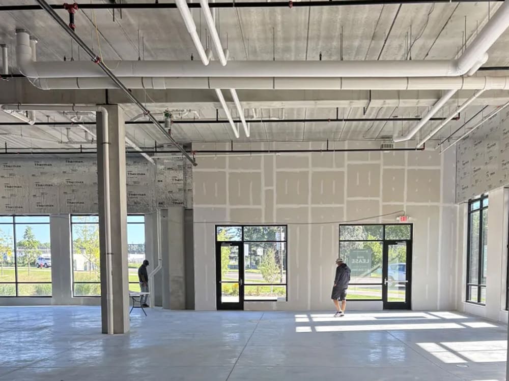A large, unfinished commercial space with concrete floors, exposed ceiling pipes, and partially completed drywall. Two people stand near the windows and glass doors, with sunlight streaming in.