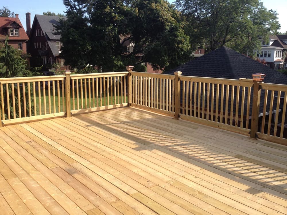 A spacious wooden deck with a railing, located outdoors and surrounded by trees and rooftops of neighboring houses on a sunny day.