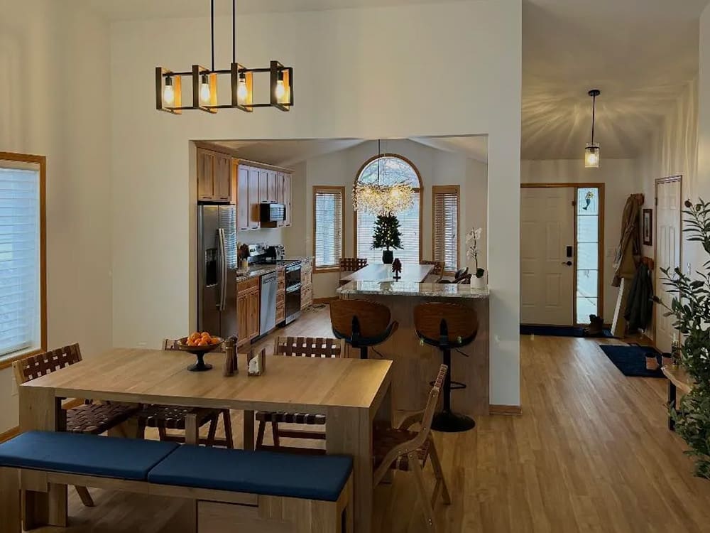 Open-concept kitchen and dining area with wooden flooring and furniture, a table with chairs and a blue bench, pendant lights, and a kitchen island with stools. The entryway is visible in the background.