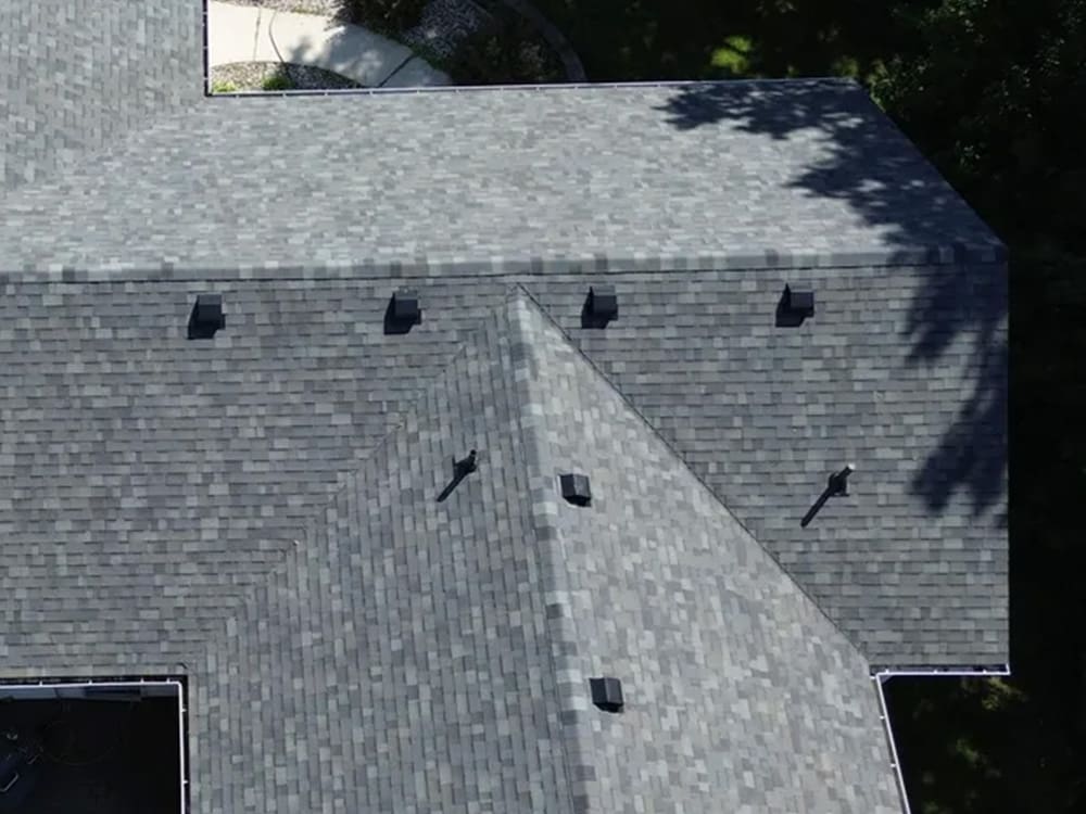 Aerial view of a house roof with gray asphalt shingles, featuring several small black roof vents. The roof has multiple angles and is surrounded by trees casting shadows.