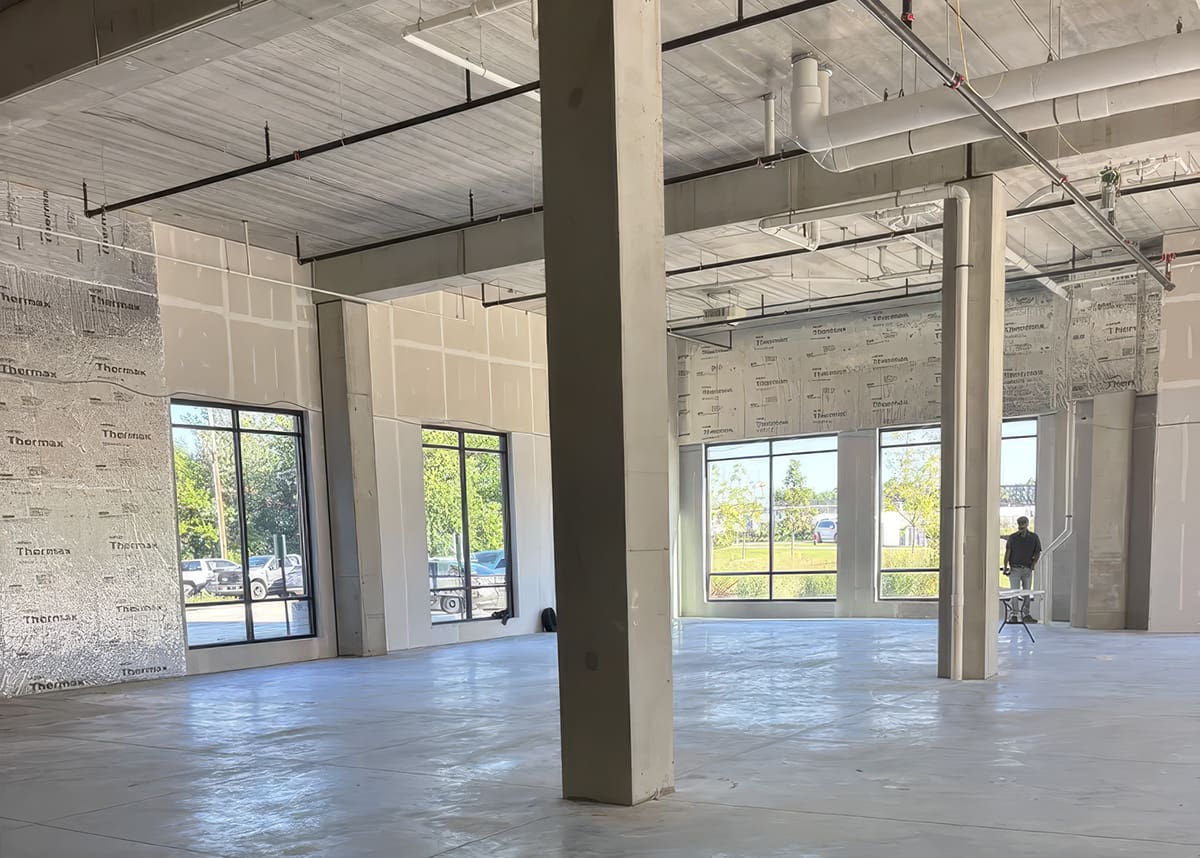 Interior of a large, unfinished commercial space with concrete floors, exposed pipes, and large windows letting in natural light; ideal for businesses seeking versatile Commercial Services. A person stands near the back by the windows.