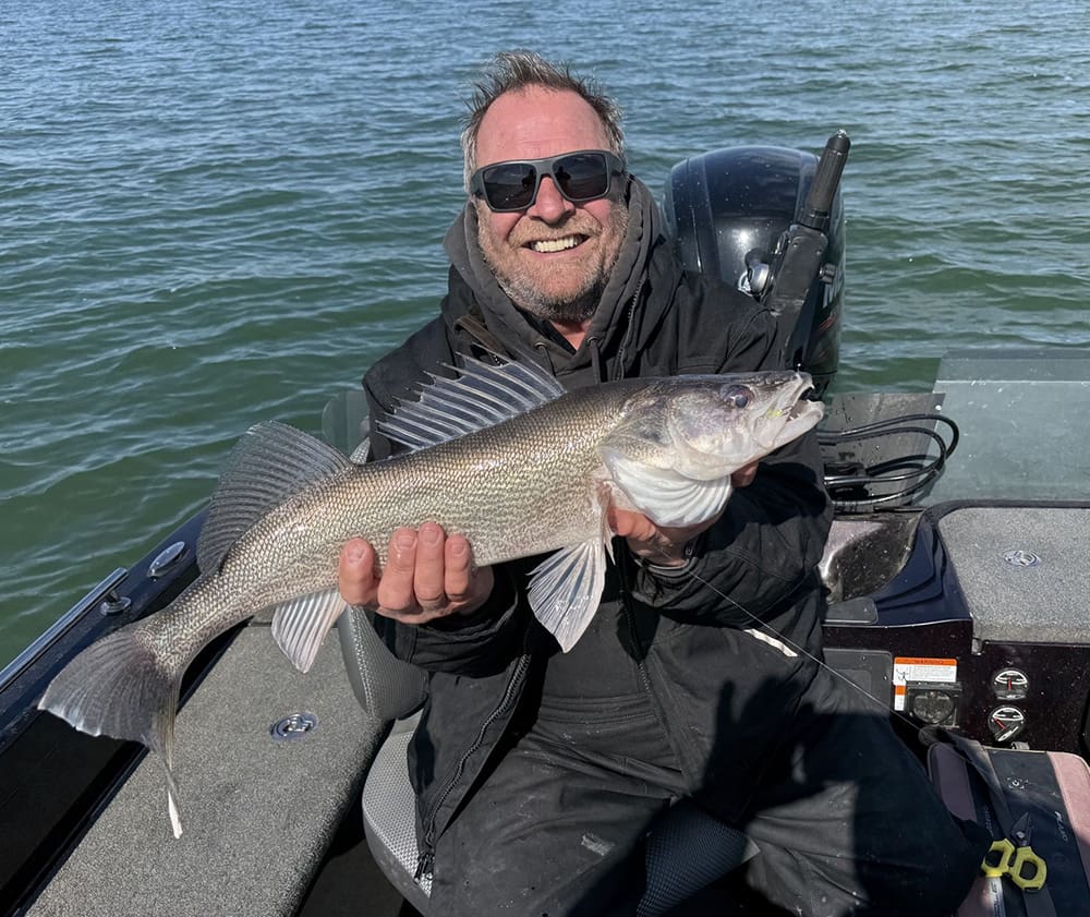 A smiling person wearing sunglasses and a dark jacket holds a large fish while sitting in a boat on the water. The sun is shining and the background shows rippling water.